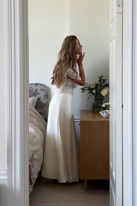 Vit linnekjol och figurnära topp bärs av en person som står nära ett trämöblemang med blommor. Neutrala färger och mjuka texturer dominerar rummet.
