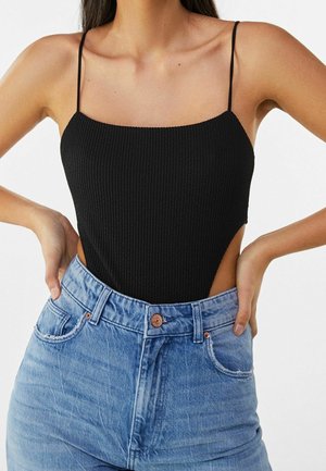 Frau trägt einen schwarzen, strukturierten, ärmellosen Body mit dünnen Trägern und hochtaillierte hellblaue Jeans, Hände auf der Hüfte ruhend.