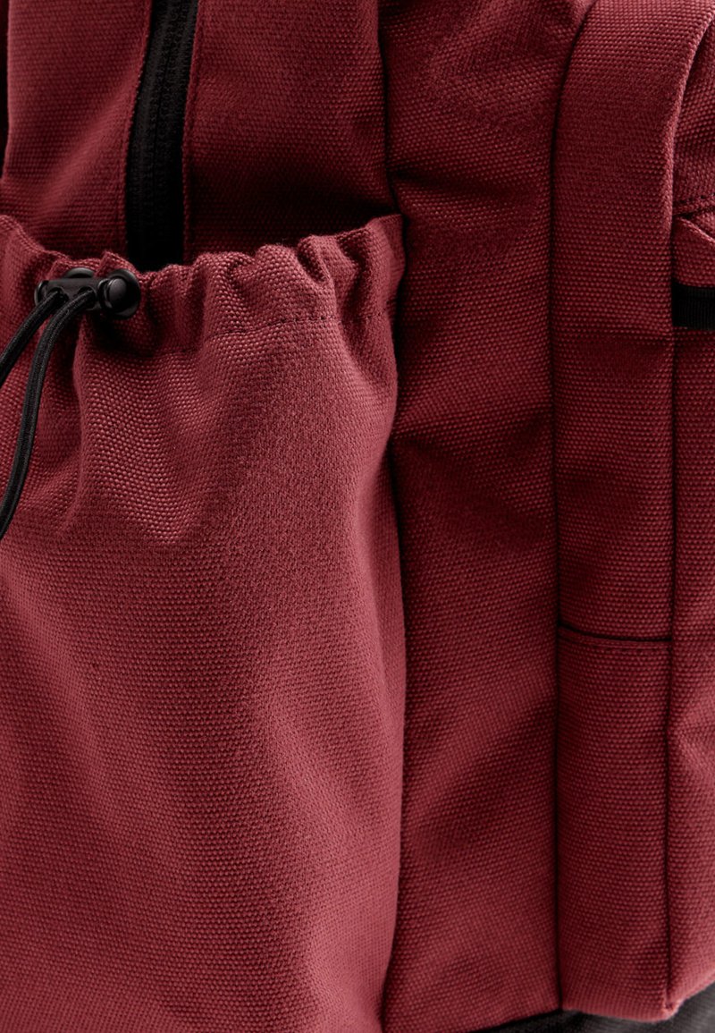 Sac à dos en tissu rouge avec un matériau texturé, une poche à cordon de serrage sur le côté et des détails en fermeture éclair noire. Design compact et structuré.