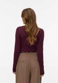 Verepunase varrukatega kampsun, mida kannavad helepruunide kõrge vöökohaga pükstega, millel on kaks tagataskut ja sile tekstuur.