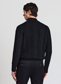 Schwarzer Strickpullover mit Fischgrätenmuster, rundem Kragen und geripptem Saum. Trägt dazu schwarze Hose, von hinten gesehen.