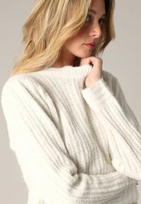 Pull en maille côtelée blanche en tissu doux, avec un col montant et une coupe décontractée, présentant une texture visible et un design confortable.