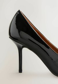 Fekete lakkbőr magas sarkú cipő, hegyes orral és vékony stiletto sarkakkal, belül barna textúrájú béléssel.