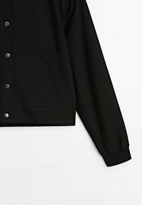 Veste noire boutonnée avec une grande poche sur le côté gauche et un poignet côtelé sur la manche droite, présentée sur fond blanc.