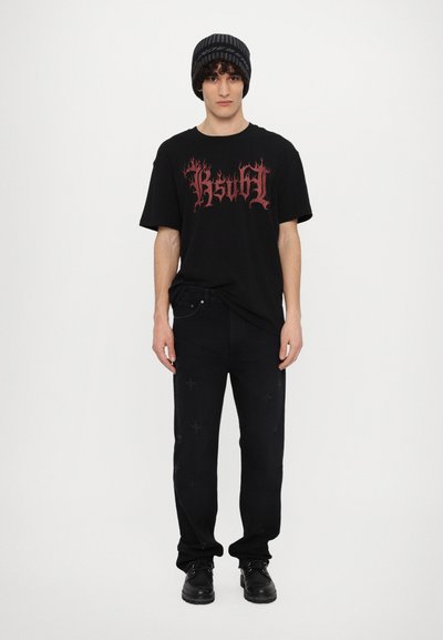 T-shirt noir surdimensionné avec un texte en flammes rouges, associé à un pantalon en denim noir présentant des motifs croisés, complété par un bonnet noir à rayures.