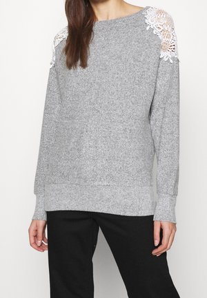 Vrouw draagt een grijze trui met lange mouwen en witte kanten uitsnijdingen op de schouders, gecombineerd met een zwarte broek, staand tegen een effen achtergrond.