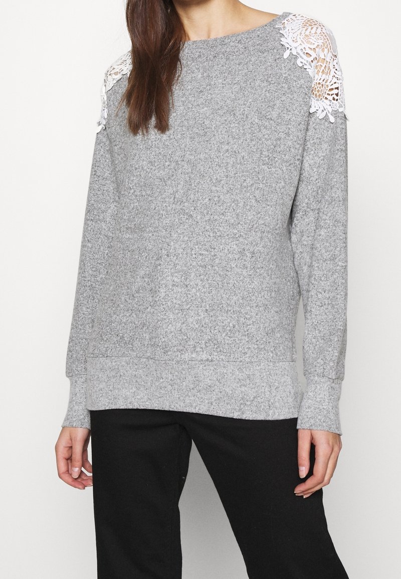 Femme portant un pull gris à manches longues avec des découpes en dentelle blanche sur les épaules, associée à un pantalon noir, se tenant devant un fond uni.