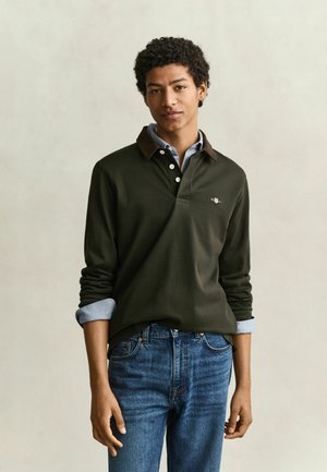 Jonge man met kort krullend haar die een donkergroene lange mouwen polo draagt over een lichtblauw overhemd en blauwe spijkerbroek, staand tegen een effen achtergrond.