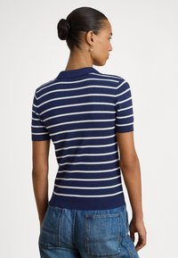 Sieviete ar matiem sasietiem bulciņā, ģērbusies tumši zilā un baltā svītrainā īsroku džemperī un zilās džinsa biksēs, skatās prom ar muguru pret balto fonu.