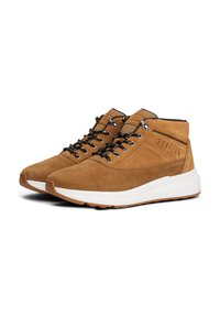 Une paire de bottes de randonnée en daim beige avec des lacets noirs et blancs, des cols rembourrés et des semelles en caoutchouc blanches, positionnées côte à côte sur un fond blanc.