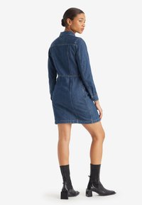 Denim-mekko, jossa on muotoiltu vyötärö, pitkät hihat ja nappikaulus. Tumma sininen kangas, jossa on ommeltuja yksityiskohtia. Yhdistetty mustiin nilkk bootseihin.