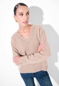 Maglione beige lavorato a maglia con scollo a V e decorazioni in perline argentate lungo il colletto, con maniche lunghe e vestibilità rilassata. Indossato con jeans blu.