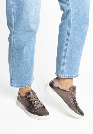 Füße in metallisch bronzefarbenen Sneakers mit weißen Sohlen und hellblauen Jeans aus Denim vor weißem Hintergrund.