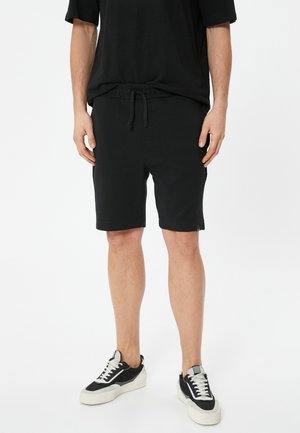 Shorts noirs avec une taille élastique et un cordon de serrage, présentant une texture lisse et une coupe jusqu'aux genoux. Associés à des baskets noires et blanches.