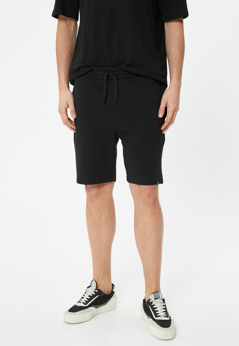 Sorte shorts med elastisk talje og snøre, med en glat tekstur og knælang længde. Sammen med sorte og hvide sneakers.