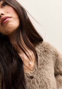 Brauner, flauschiger Cardigan mit einem Knopf, geschichtete goldene Halsketten und eine Perlenakzent. Das Model hat lange dunkle Haare und ist im Winkel positioniert.