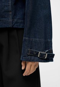 Dunkelblaues Jeansjackenärmel mit einer metallischen Schnallen-Detail und kontrastierenden Nähten; glatte Textur und taillierter Schnitt am Bündchen.
