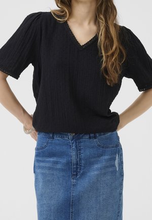 Femme portant un haut noir texturé à manches courtes en V et une jupe en denim bleue, les mains posées sur les hanches, cheveux longs et ondulés tombant sur l'épaule.