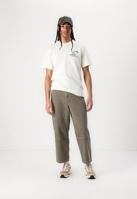 Kremfarget t-skjorte med korte ermer, grafikk og rød tekst, olivengrønne baggy bukser med sidesømmer, og kremfargede sneakers med svarte detaljer.
