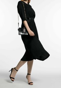 Svart knälång klänning med en åtdragen midja, kombinerad med svarta sandaler med remmar. Modellen håller en texturerad svart och silverhandväska.
