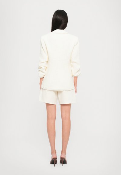 Witte textuur blazer met opgerolde mouwen, gecombineerd met bijpassende shorts, beide in een lichte stof. Draagt zwarte hoge hakken.