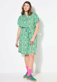 Abito verde a fantasia con maniche corte, vita legata e design circolari in tonalità di rosa e verde. Abbinato a scarpe verdi e calzini rosa.