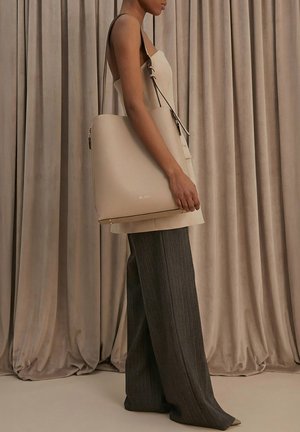 Žena v béžovém tílku bez rukávů a šedých širokých kalhotách s tenkými proužky, držící velkou béžovou koženou tašku přes rameno.