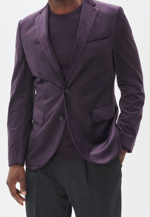 Homme portant un blazer en velours violet foncé et un pull assorti avec un pantalon noir, une main dans la poche, sur un fond clair uni.