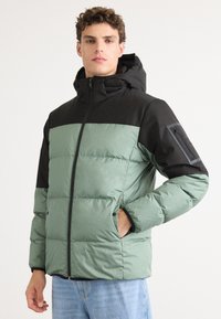 Groene pufferjack met een zwart bovenstuk en capuchon. Beschikt over een ritszak op de linkermouw en twee zijzakken. Glad, geïsoleerd textuur.