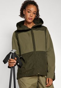 Olivenfarget, vanntett jakke med hette, med glidelås og trykknapp-lukking, og et to-farget design. Holdt med turstaver.
