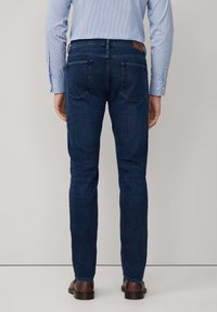 Muškarac nosi tamnoplave uske traperice, svijetloplavu prugastu košulju i smeđe kožne cipele, stoji naslonjen na svijetlu jednobojnu pozadinu.