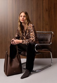Jeune femme en blouse à imprimé léopard et pantalon noir assise sur une chaise moderne, tenant un grand sac à main marron devant un fond en panneaux de bois.