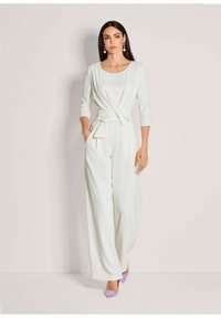 Witte jumpsuit met driekwartmouwen, een strikceintuur, een brede broekspijp en zakken. Gecombineerd met lavendel hoge hakken en opvallende oorbellen.