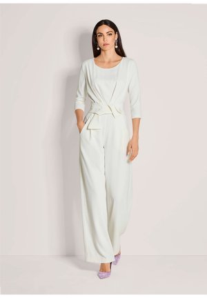White jumpsuit with three-quarter sleeves, a tie waist, wide leg, and pockets. Paired with lavender high heels and bold earrings.