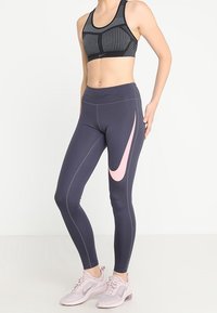 Sujeción deportiva de patrón negro y gris, leggings de alta cintura en gris oscuro con un gran logotipo de Nike en color rosa, y zapatillas deportivas en rosa claro.