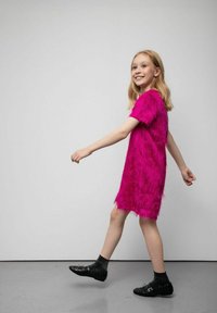 Jaskrawy różowy, puszysty sukienka z krótkimi rękawami i ozdobnymi frędzlami, do kolan, noszona z czarnymi skarpetkami i butami, na jednolitym tle.