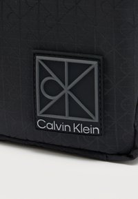 Μαύρη τσάντα με υφή καπιτονέ, με ένα εμφανές γκρι λογότυπο με γεωμετρικό σχεδιασμό και την επιγραφή «Calvin Klein» στο κάτω μέρος.