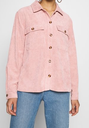 Mujer con una camisa de pana rosa con botones y dos bolsillos en el pecho, combinada con jeans azules.