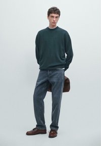 Tmavě zelený pletený svetr, volný střih. Světle šedé džíny, mírně volné. Hnědé kožené boty. Model drží hnědou tašku, neutrální pozadí.
