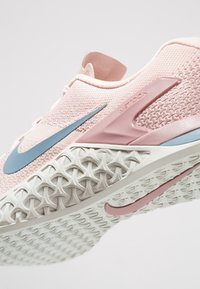 Zapatilla deportiva rosa con una parte superior de malla texturizada, que presenta un swoosh azul y una suela de goma blanca con un patrón en el dibujo para mayor agarre.