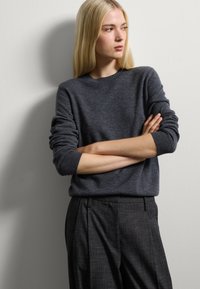 Pull en laine gris foncé avec des manches retroussées, associé à un pantalon noir texturé. Le modèle pose contre un mur clair, les bras croisés.