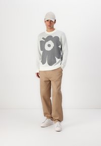 Helles graues Sweatshirt mit großem dunkelgrauen Blumenmuster, kombiniert mit beigen Hosen und weißen Sneakers. Ergänzendes hellbeiges Cap.