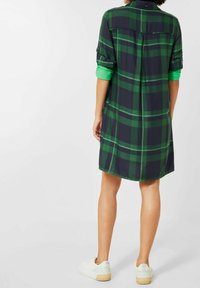Donna che indossa un vestito camicia a quadri verde e blu navy con maniche arrotolate e sneakers bianche, in piedi contro uno sfondo grigio chiaro uniforme.