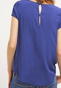 Blusa a maniche corte blu reale con dettaglio a oblò sul retro e chiusura con bottone, realizzata in tessuto liscio con vestibilità rilassata e orlo curvo.
