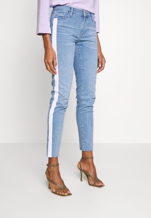 Hellblaue Skinny-Jeans mit weißen Seitenstreifen, kombiniert mit olivgrünen Riemchen-Sandaletten mit Absatz und einem lavendelfarbenen Langarmtop.