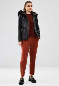 Giacca di pelle nera con cappuccio in pelliccia sintetica, abbinata a pantaloni sartoriali color ruggine e scarpe con plateau nere con dettagli dorati.