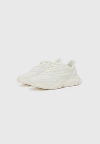 White sneaker featuring a smooth synthetic upper, lace-up closure, rounded toe, and a thick rubber sole with subtle textured details.