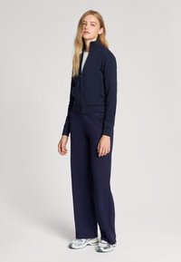 Giacca a zip blu marine con colletto alto, abbinata a pantaloni a gamba larga coordinati. Modello che indossa sneakers grigie. Texture liscia, dettagli di design minimali.