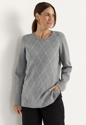 Vrouw met bruin haar in een staart, draagt een grijze trui met lange mouwen en een wit ruitpatroon en zwarte broek, staand tegen een witte muur.