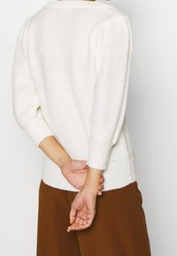 Pull en maille blanche à coupe ample, design texturé et poignets côtelés. Présente un large col et des manches légèrement bouffantes.